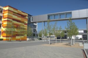 Gelb-Orange-Rotes Gebäude mit Grauer Gebäudeüberbrückung über dem Schulhof